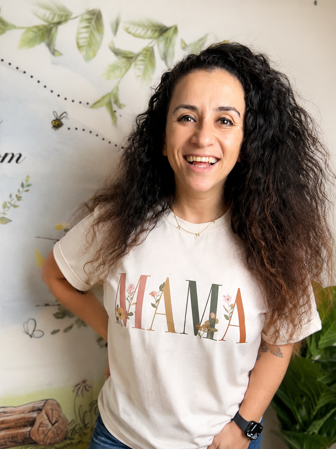 Fate Winczuk smiling in a MAMA t-shirt against a hand-painted nature wall